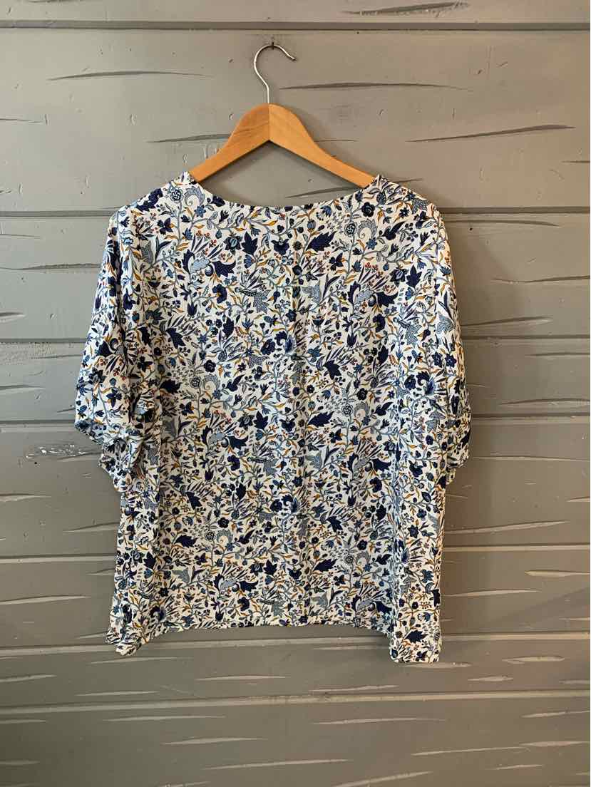 White blouse with blue and black floral pattern, three-quarter sleeves, round neckline, hanging on wooden hanger.