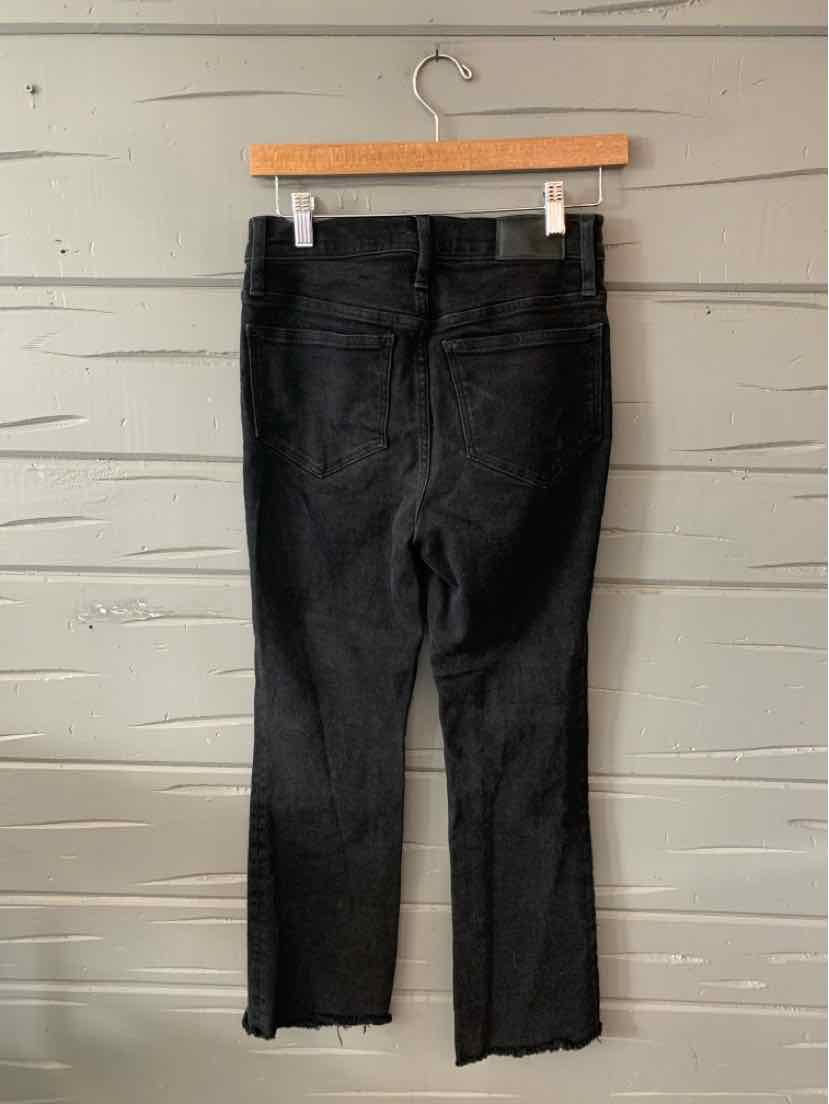 Black denim jeans with a straight leg and raw hem hanging on a wooden hanger against a light wood wall.