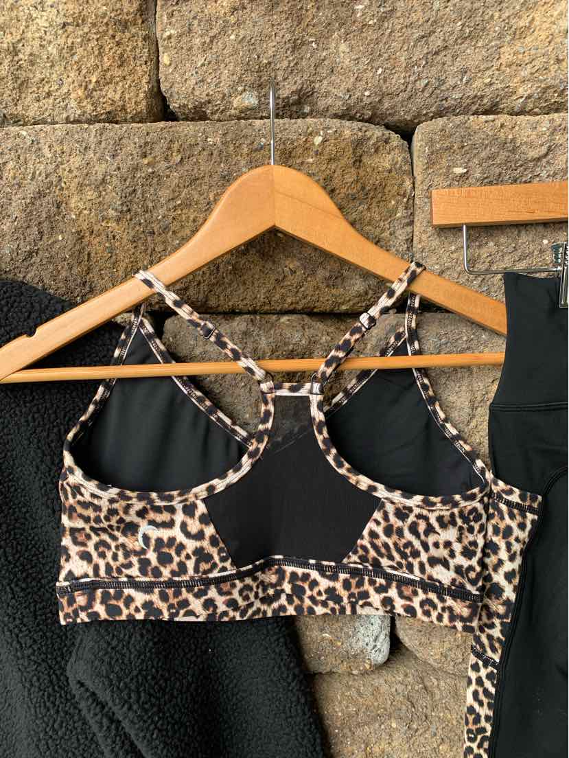 Leopard print sports bra with black back panel and crisscross straps on wooden hanger against stone wall.