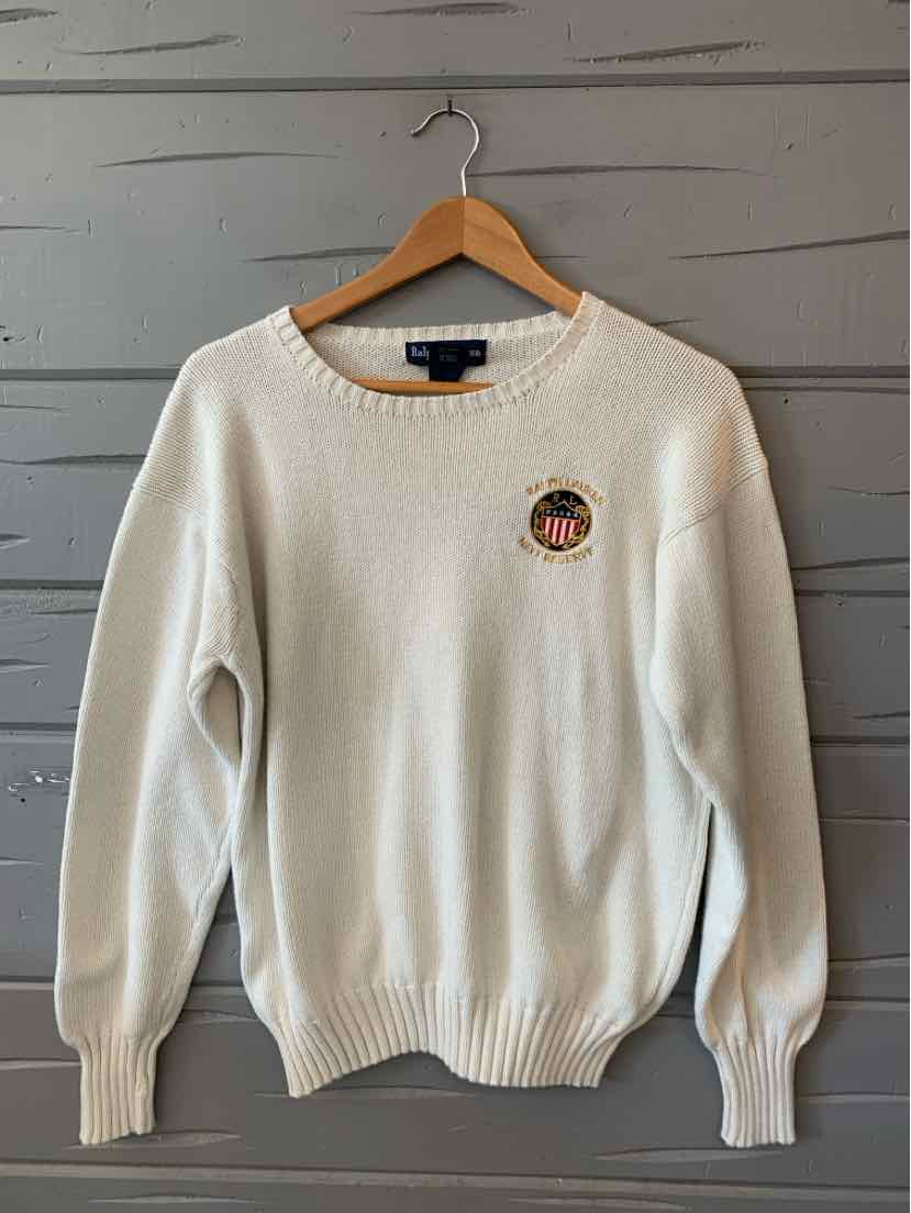 Cream-colored knit sweater with crewneck and ribbed cuffs featuring a small circular embroidered logo on the chest.