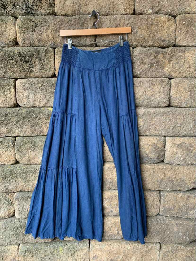 Blue wide-leg pants with tiered ruffle design and elastic waistband hanging against stone wall.