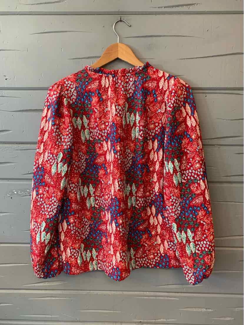 Red blouse with long sleeves and floral pattern in white, blue, and purple hanging on wooden hanger against gray wooden wall.