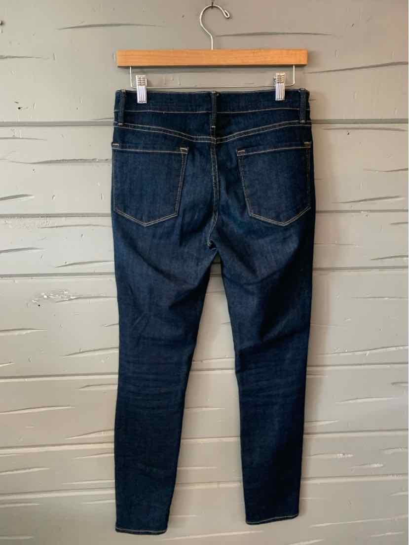 Dark blue slim-fit denim jeans with rear pockets and yellow stitching on wooden hanger against gray wooden background.