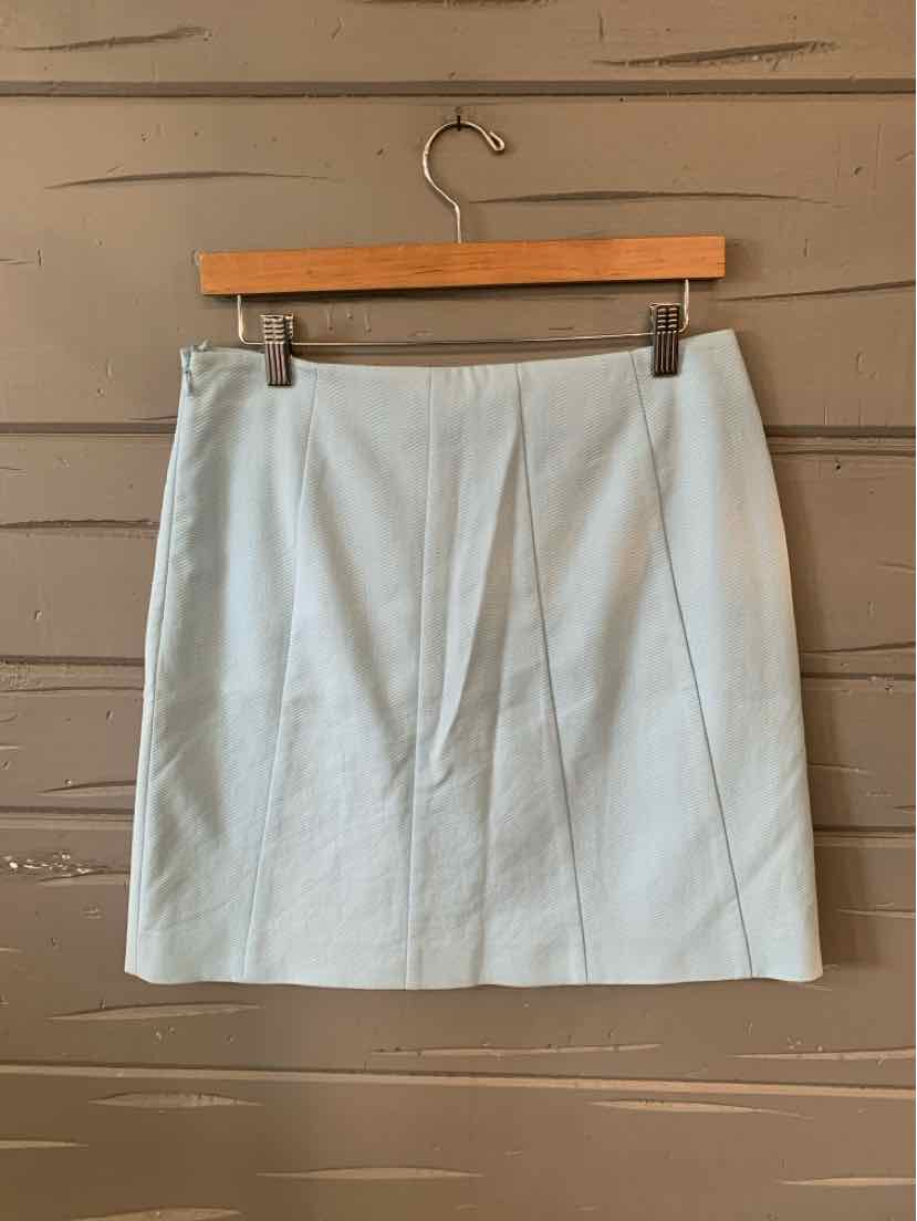 Light blue knee-length skirt with front pleats hanging on wooden hanger against tan wooden wall.