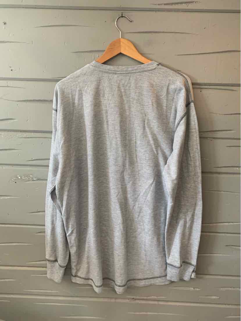 Light gray long-sleeve shirt with a crew neck, displayed on a wooden hanger against a wooden wall.