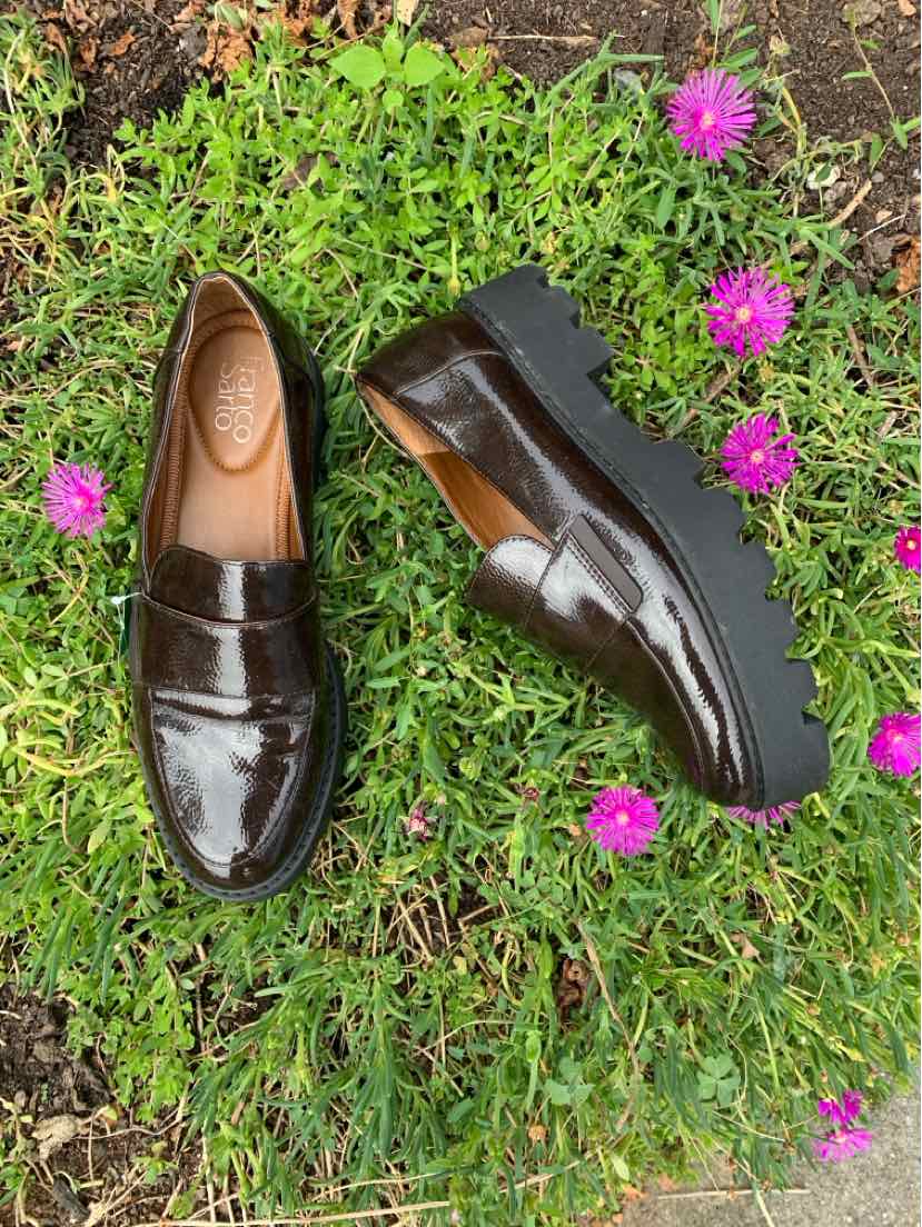 Pair of dark brown patent leather loafers with a rounded toe and low heel on green grass with pink flowers.