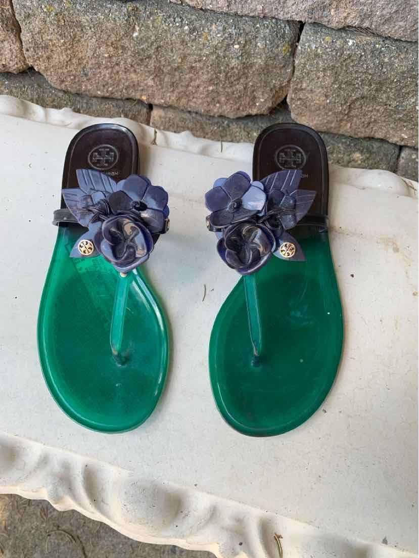 Green flat sandals with black thong straps and large decorative black flowers on top.