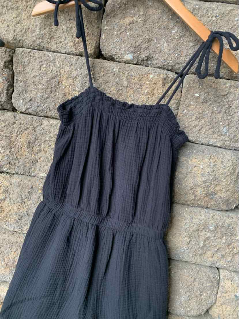 Black textured sleeveless dress with adjustable spaghetti straps and smocked bodice hanging on wooden hanger against stone.