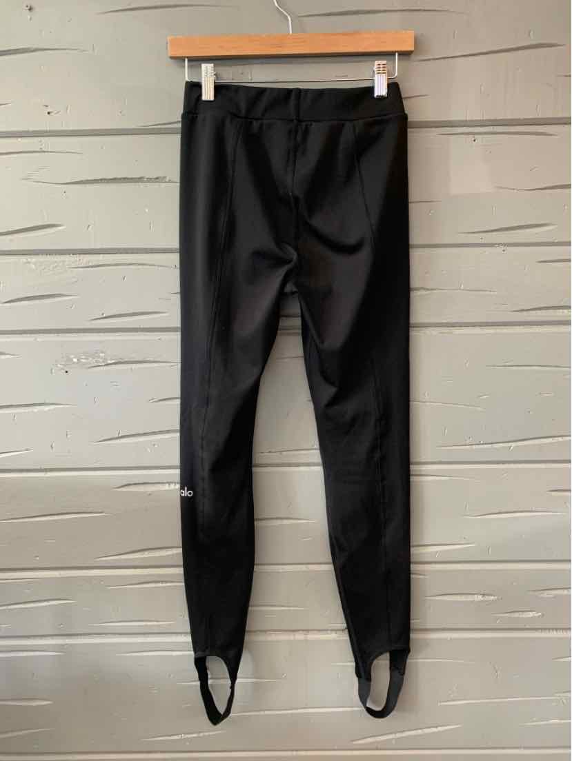 Black small women's leggings with stirrups hanging on wooden hanger against gray wall.