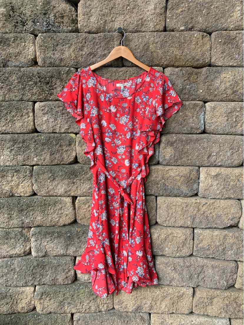 Red floral wrap dress with ruffled short sleeves and tie waist hanging on wooden hanger against stone wall.