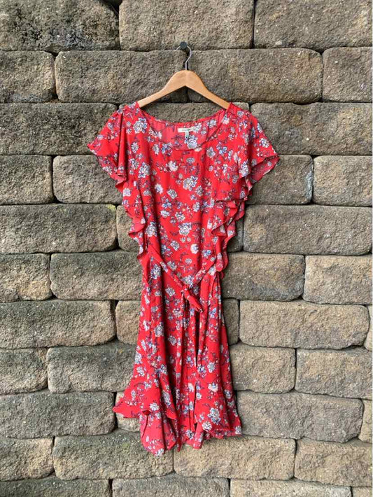 Red floral wrap dress with ruffled short sleeves and tie waist hanging on wooden hanger against stone wall.