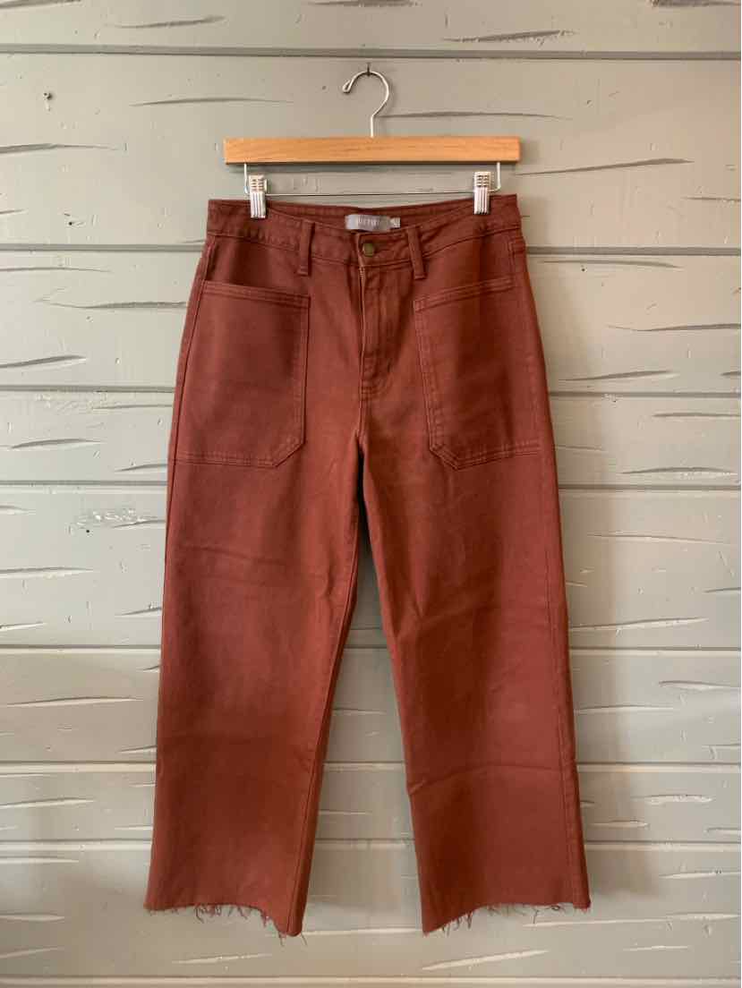 Rust-colored wide-leg pants with front patch pockets and raw hem on a wooden hanger.