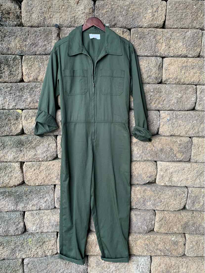 Olive green long-sleeve utility jumpsuit with front pockets and a collar, displayed hanging against a stone wall background.