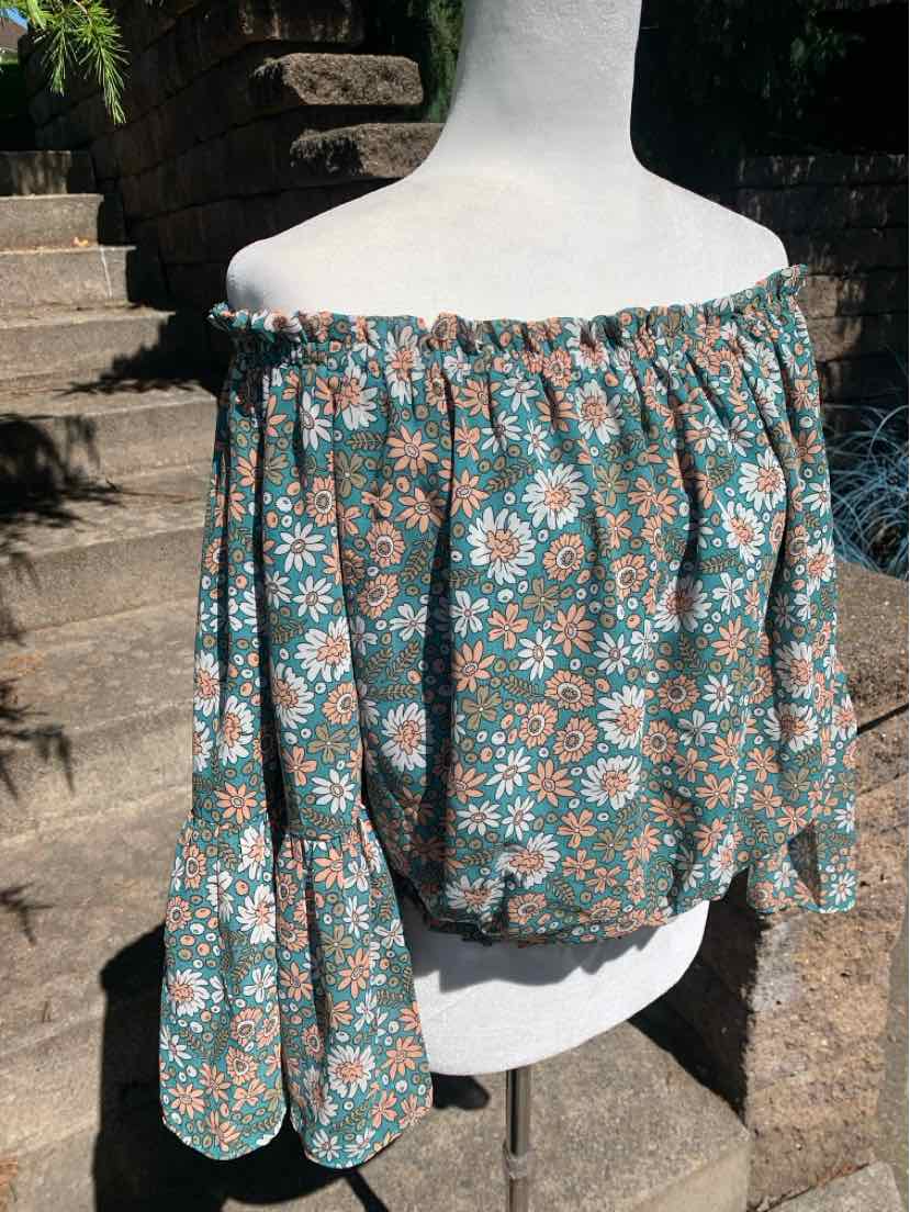 Off-shoulder blouse with elastic neckline, green base, pink and white floral print, loose long sleeves.