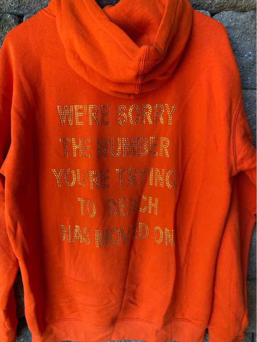 Bright orange hoodie with gold metallic text on the back reading "WE'RE SORRY THE NUMBER YOU'RE TRYING TO REACH HAS MOVED.