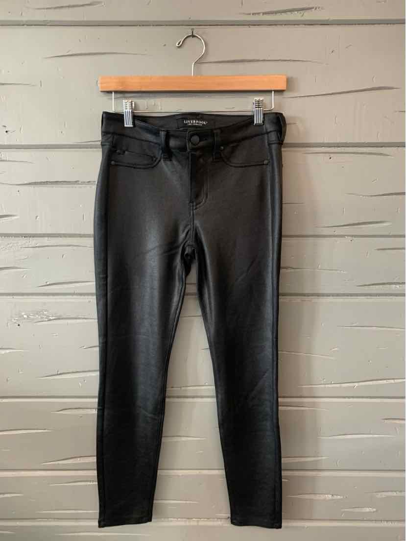 Black slim-fit pants with front pockets and button closure hanging on wooden hanger against gray wooden wall.
