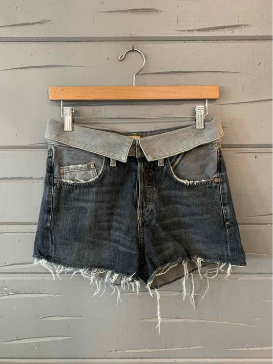 Gray denim shorts with frayed hems and front pockets hanging on wooden hanger against wooden wall.
