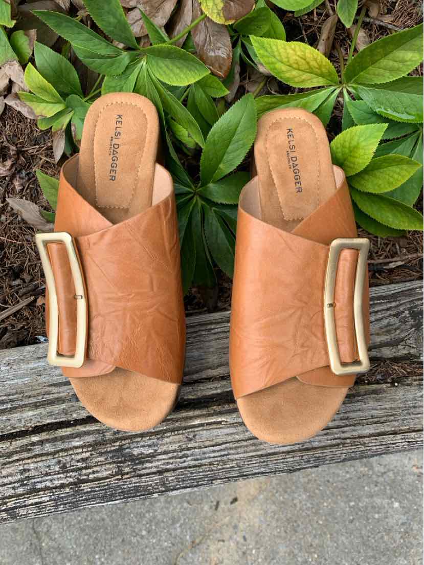 Tan leather open-toe slide sandals with wide straps and large rectangular metal buckle embellishments.
