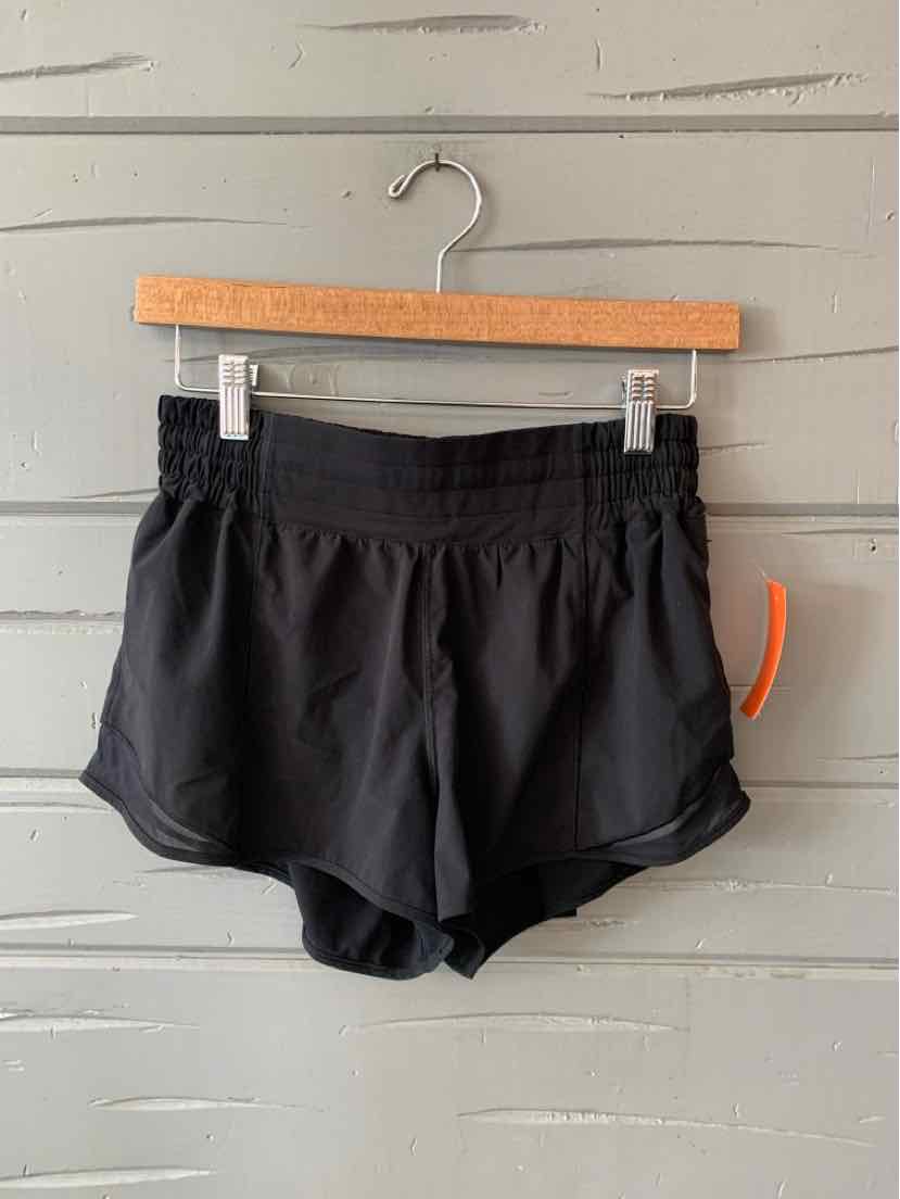 Black athletic shorts with elastic waistband and curved hems hanging on wooden hanger against gray wood panel background.