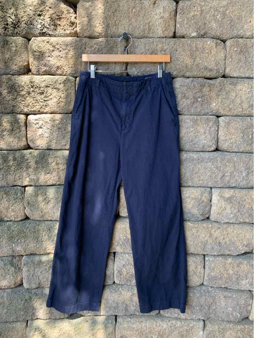 Navy blue straight-leg pants with elastic waistband and side pockets hanging on wooden hanger against stone wall.