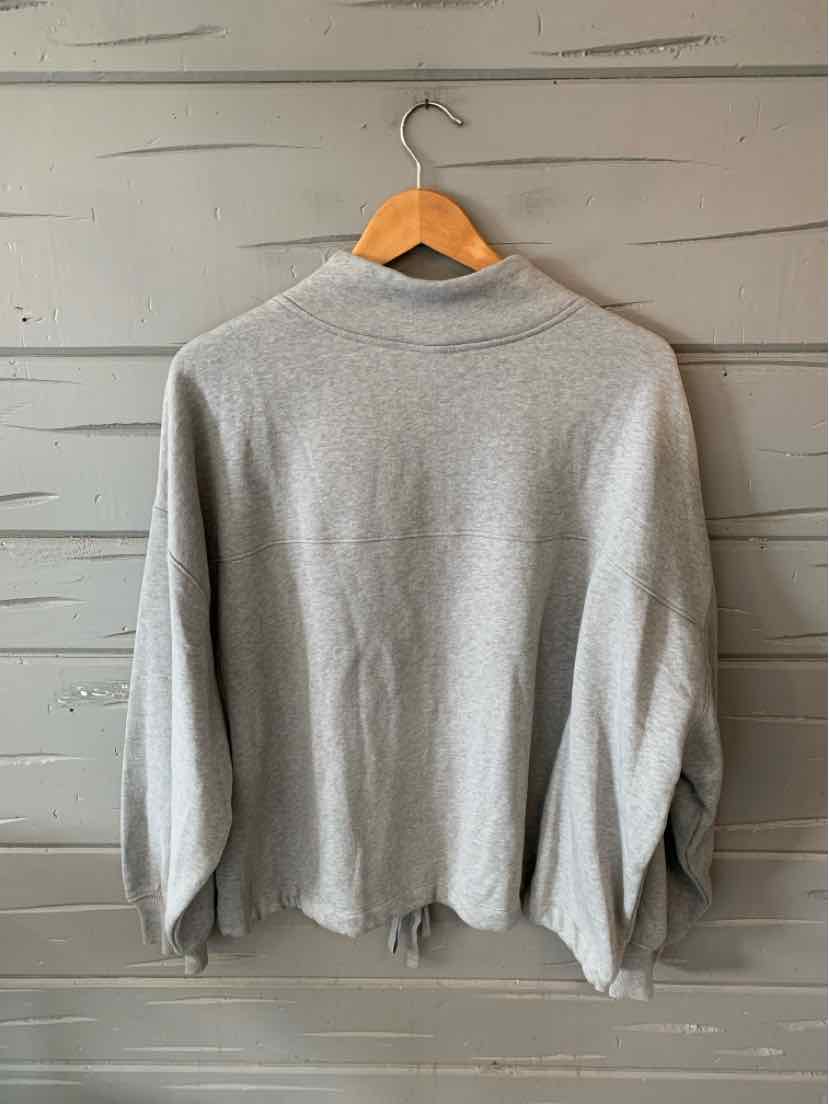 Light gray long-sleeve sweatshirt with a high collar and relaxed fit hanging on a wooden hanger against a gray wood-paneled.