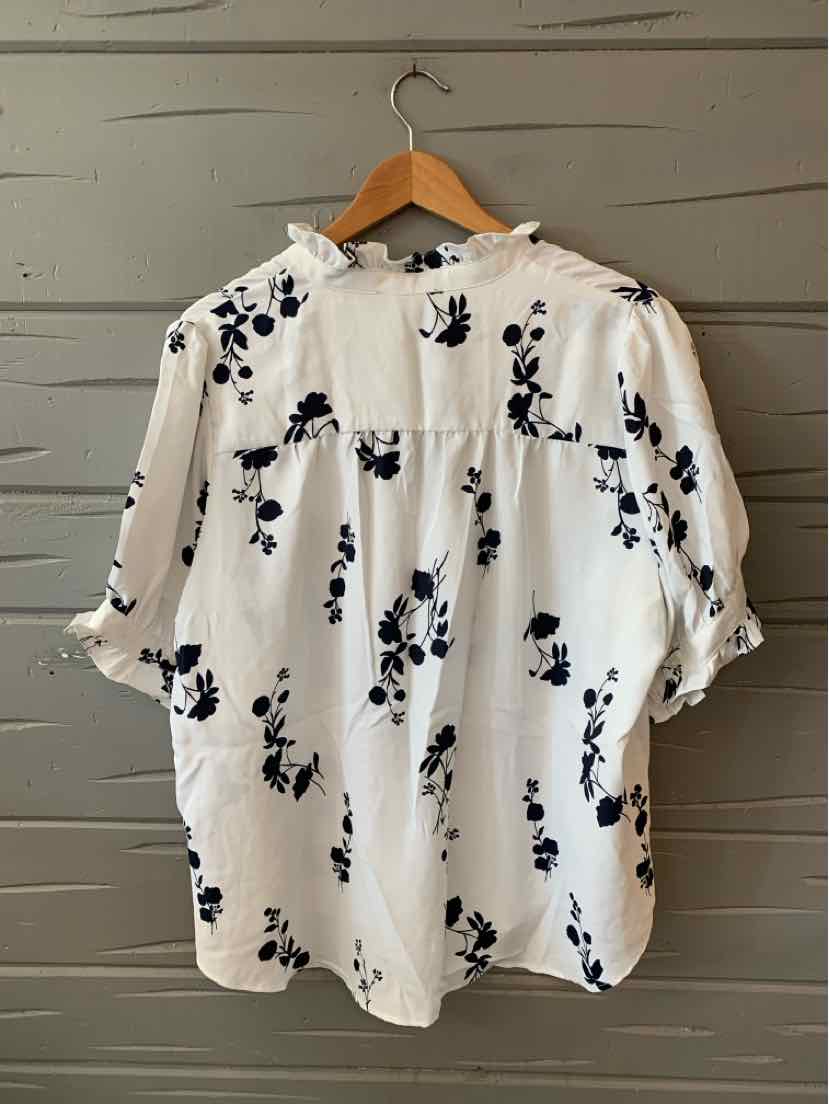 White blouse with black floral print, half sleeves, buttoned back, and loose fit on wooden hanger.