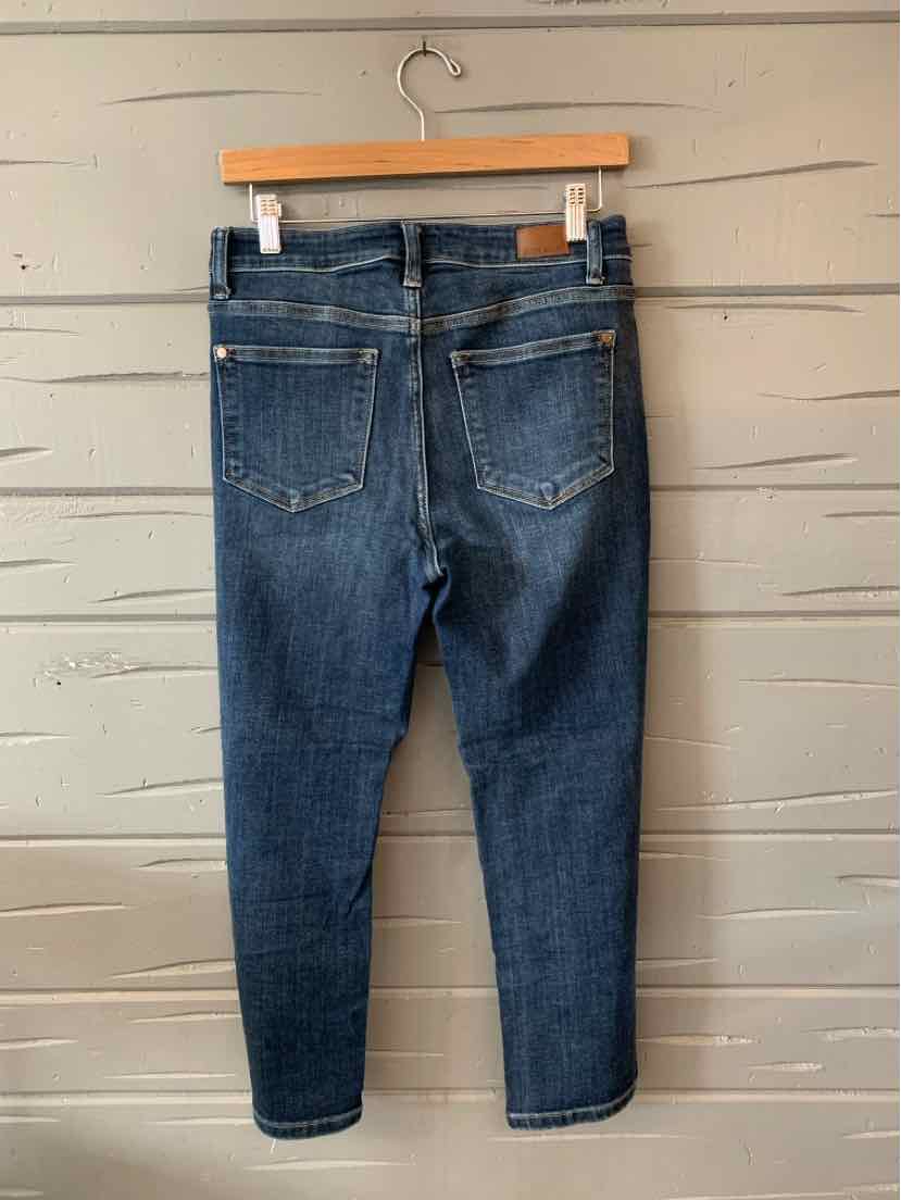 Dark blue denim jeans with back pockets and a slim, tapered leg hanging on a wooden hanger against a gray wooden wall.