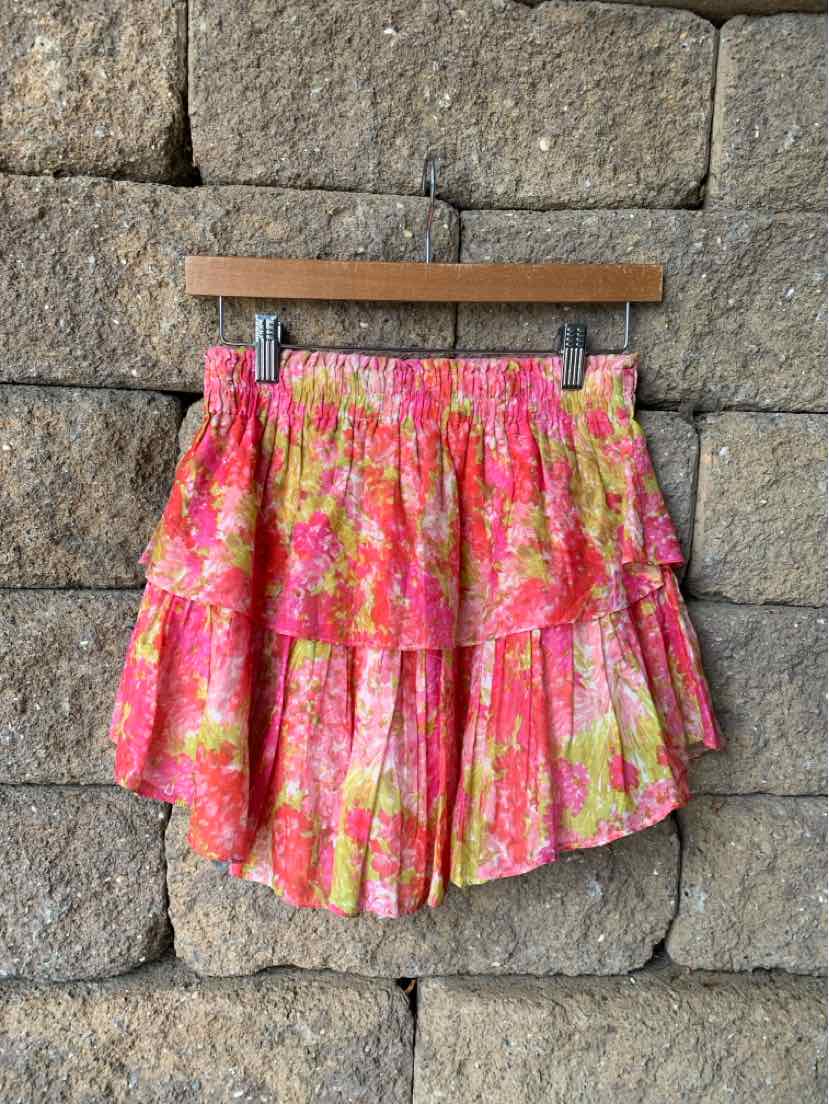 Pink and yellow floral ruffled mini skirt with an elastic waistband hanging on wooden hanger against stone wall.