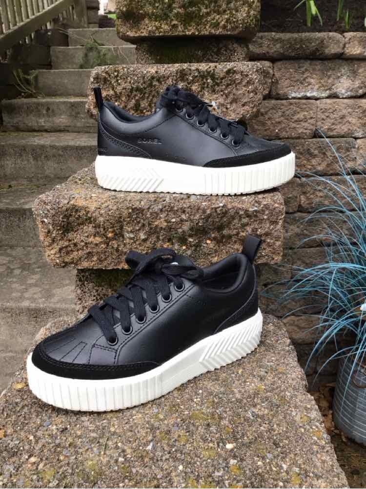 Black leather sneakers with white textured platform soles and black laces on outdoor stone steps.