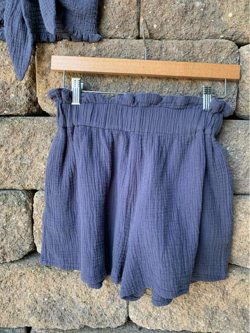Navy blue textured fabric shorts with elastic waistband and ruffled trim hanging on wooden hanger against stone wall.