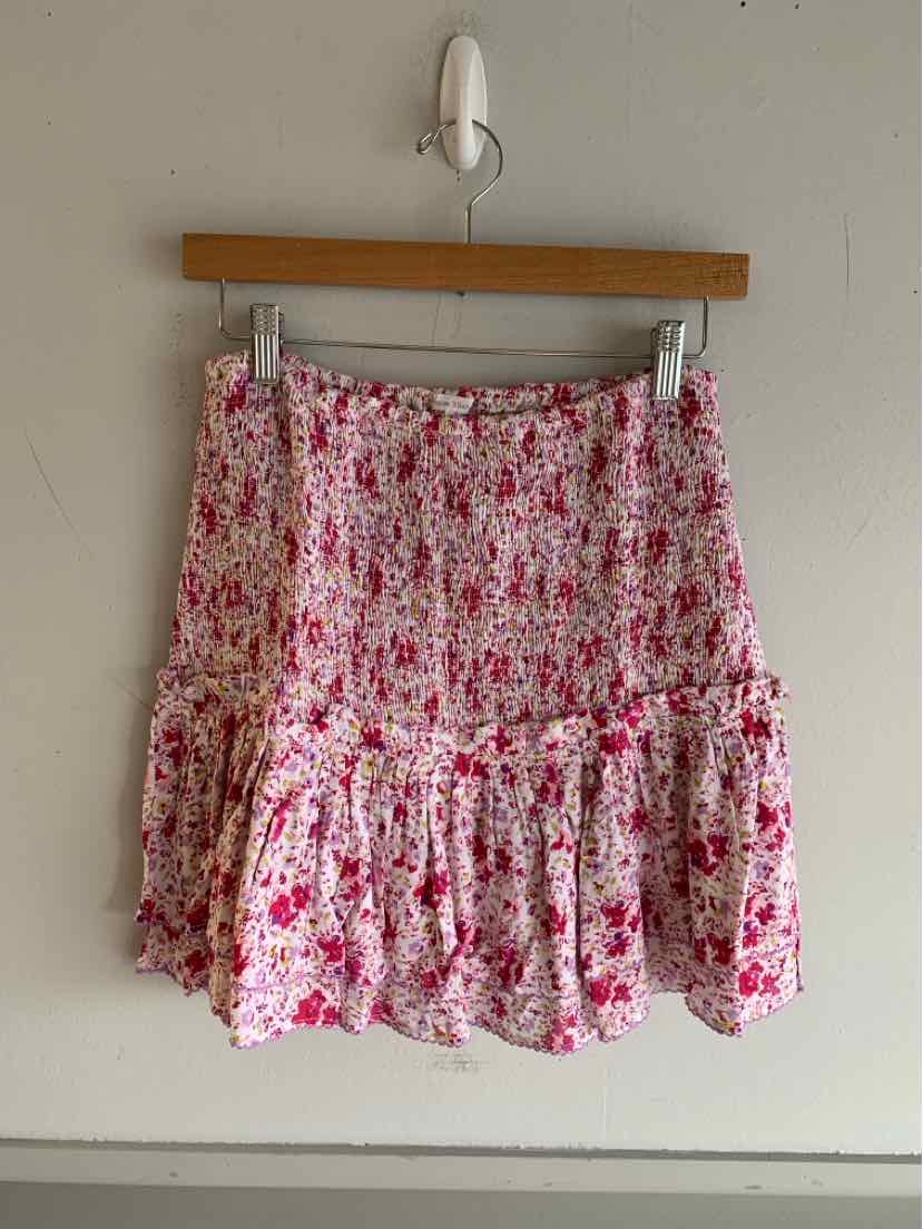 Pink floral mini skirt with ruffled hem and smocked waistband hanging on wooden hanger against beige wall.