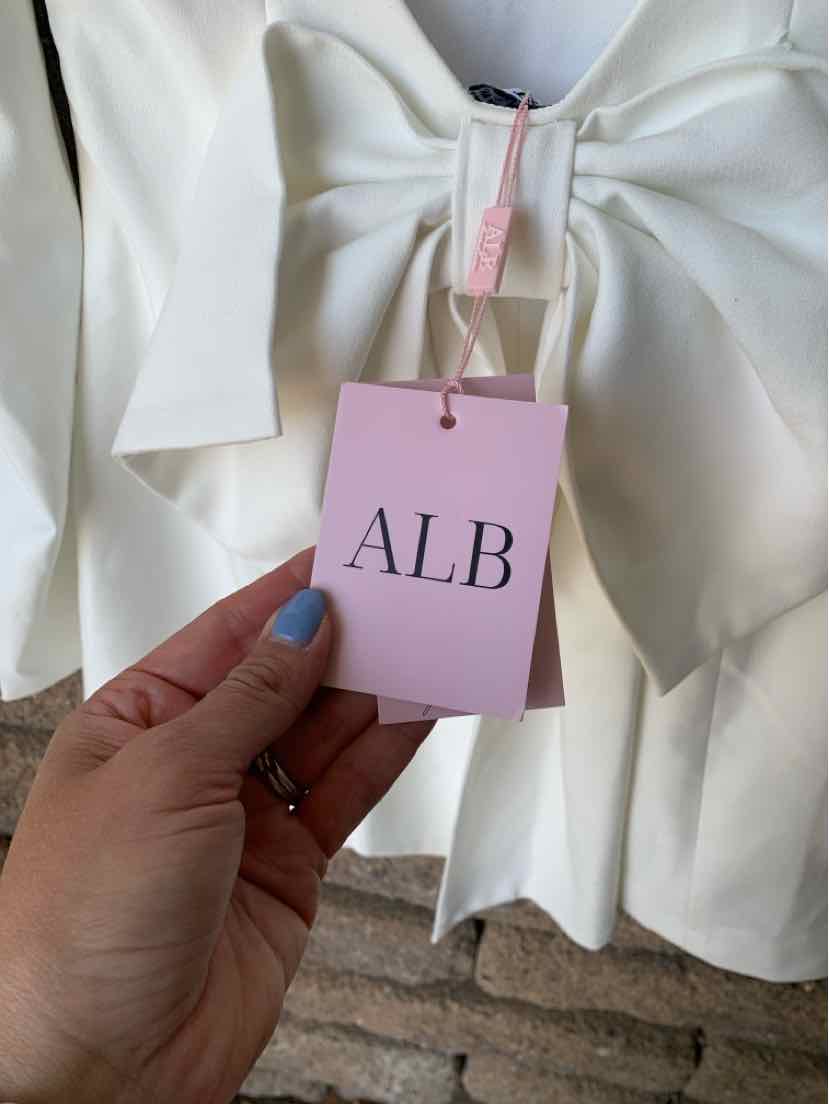 White garment with large bow detail at neckline and pink ALB tag attached.