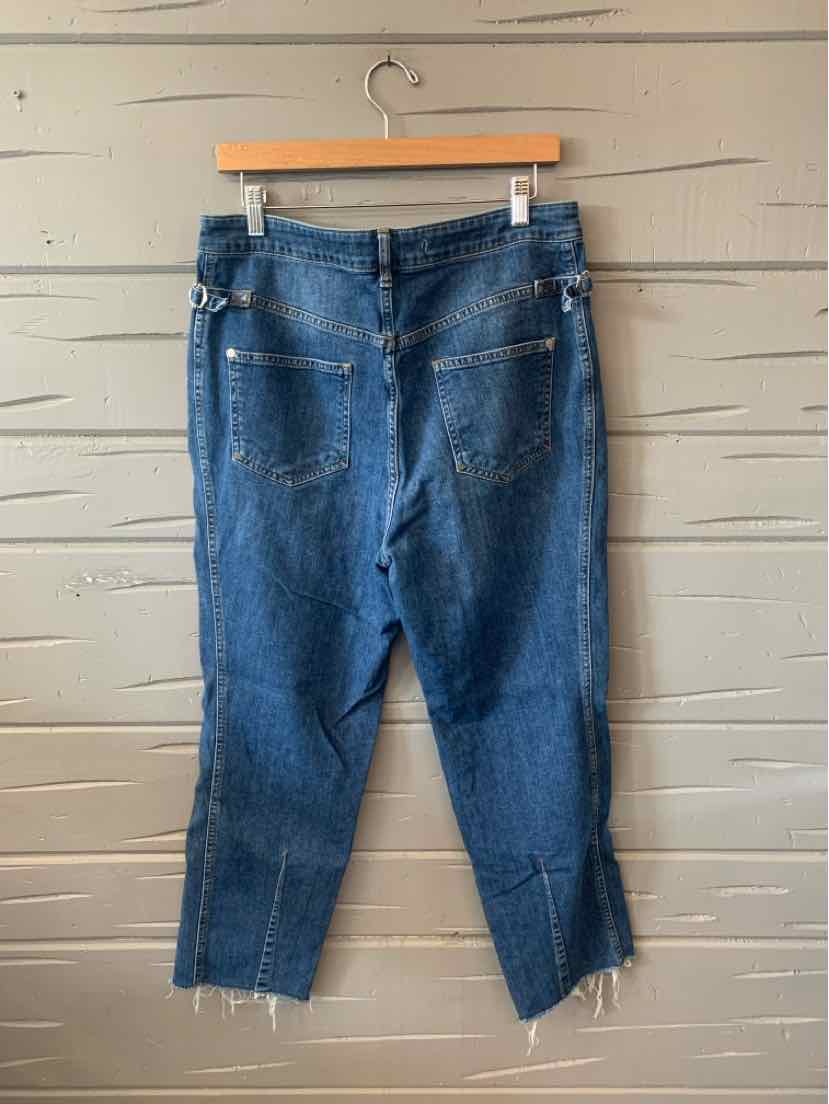 Blue cropped denim jeans with raw hems and front pockets hanging on wooden hanger against wooden wall.