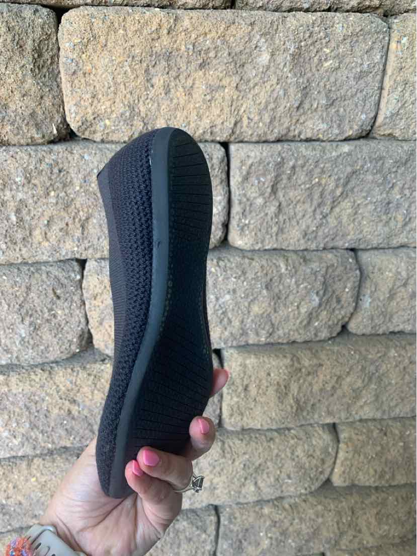 Black Allbirds shoe sole held against a beige stone wall background, size 6.5 visible in hand for scale.