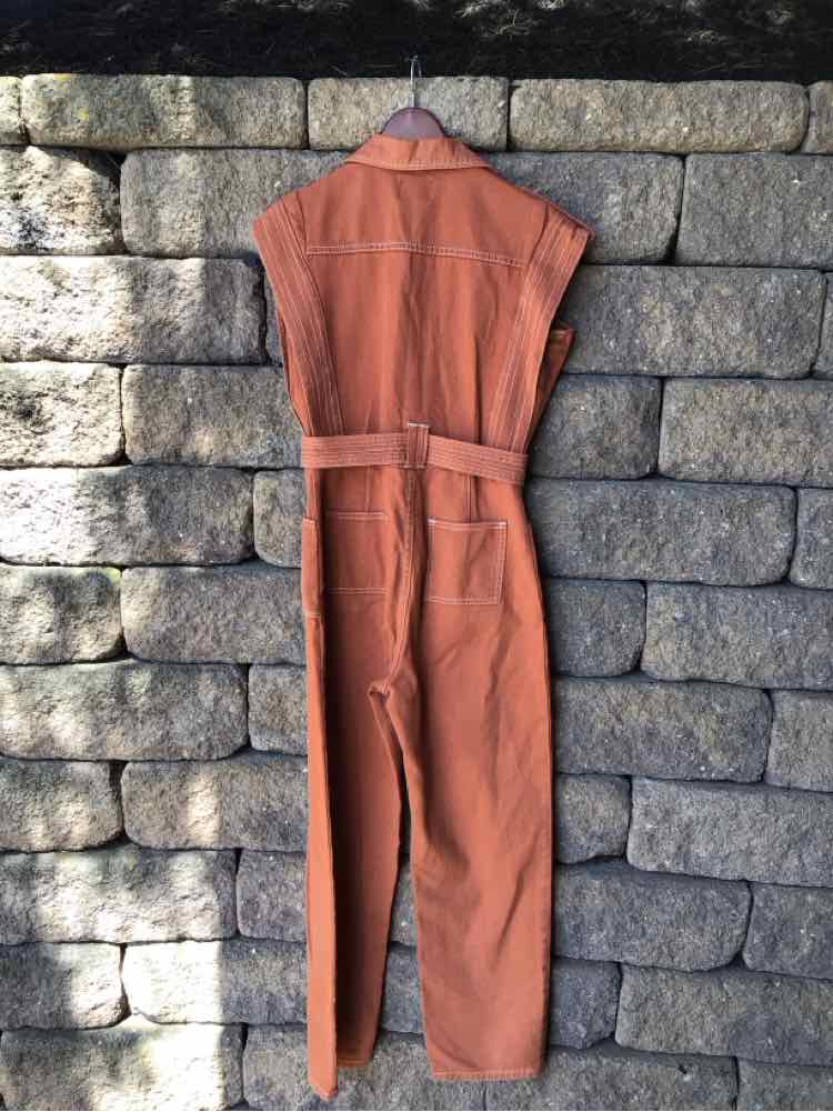 Rust orange sleeveless denim jumpsuit with belt, fitted waist, and straight legs hanging on stone wall.