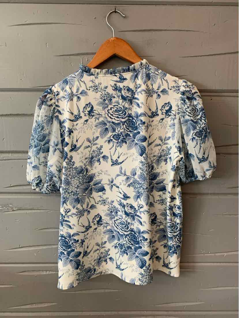 White blouse with blue floral print, puffed short sleeves, and round neckline, hanging on wooden hanger.