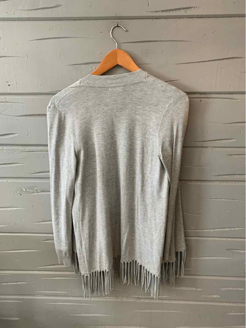 Light gray long-sleeve cardigan with fringe hem hanging on wooden hanger against gray wood wall.