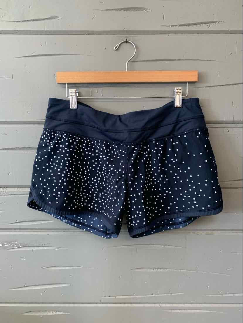 Black athletic shorts with white polka dots, wide waistband, and curved hem hanging on wooden hanger against gray wooden.