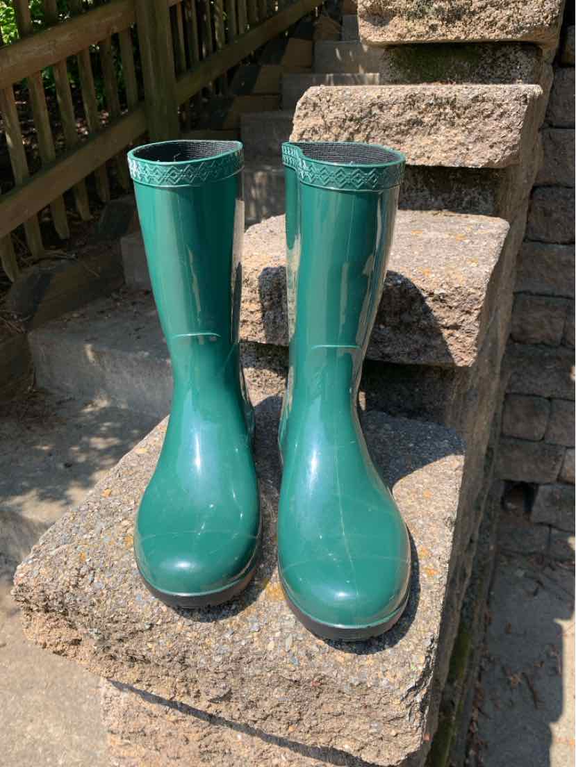 Glossy dark green knee-high rain boots with a rounded toe and lined interior standing on outdoor stone steps.