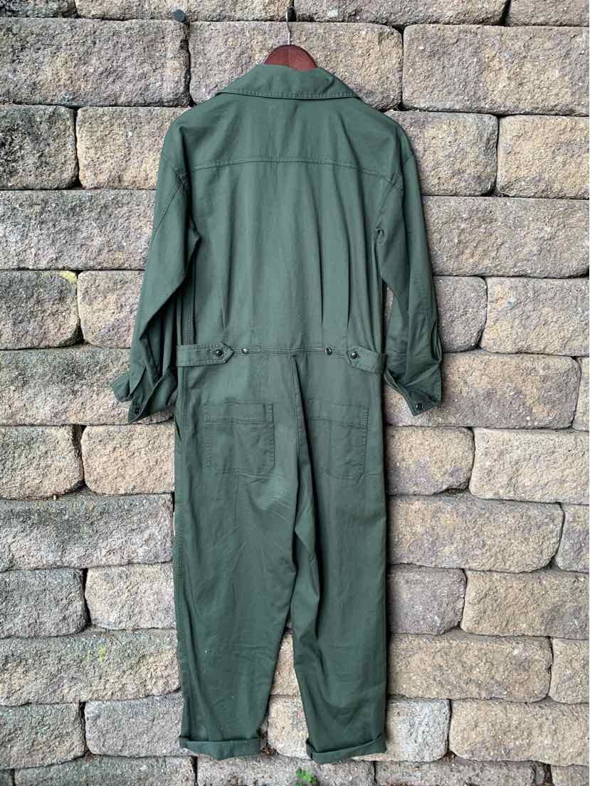 Dark green long-sleeve utility jumpsuit with cinched waist and cuffed ankles hanging against stone wall.