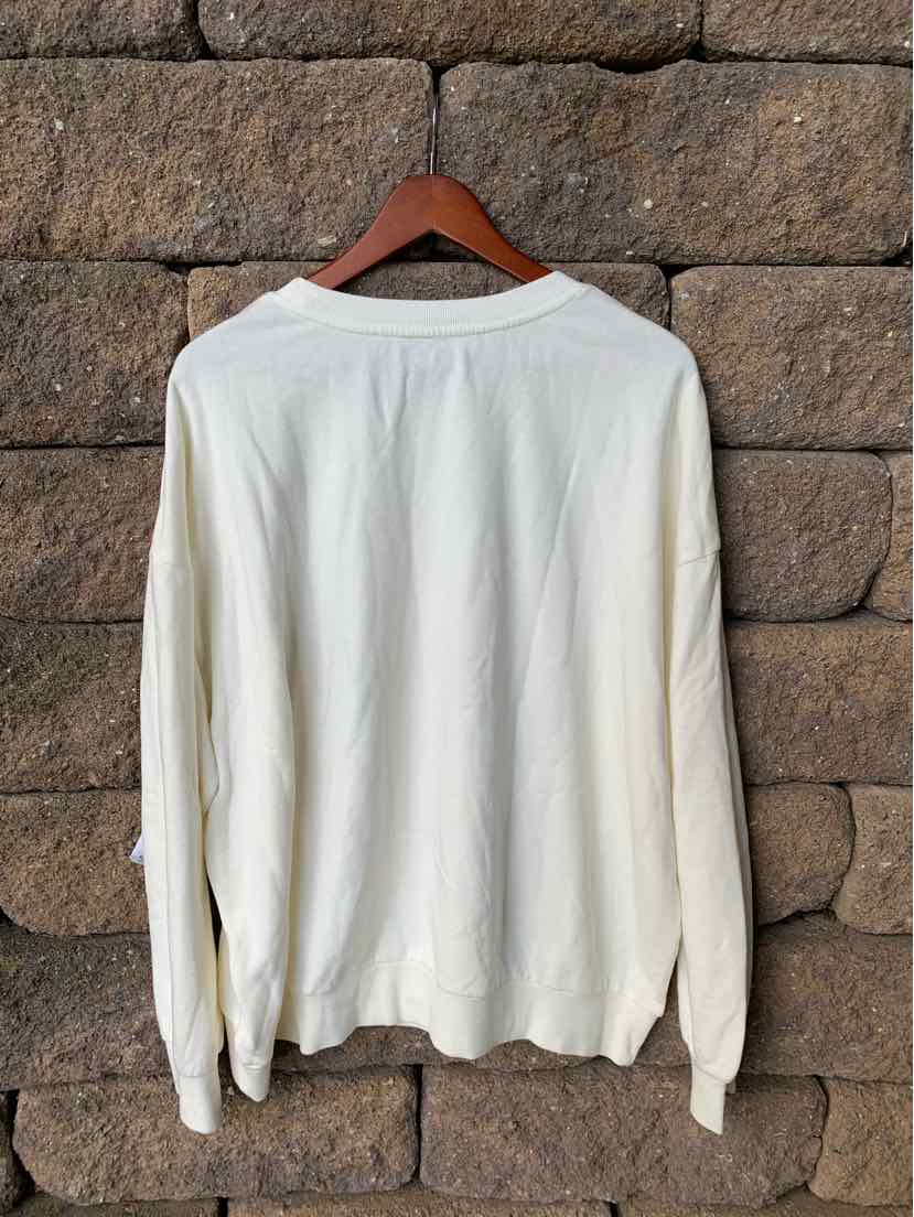 White long-sleeve crewneck T-shirt with relaxed fit on wooden hanger against stone wall.