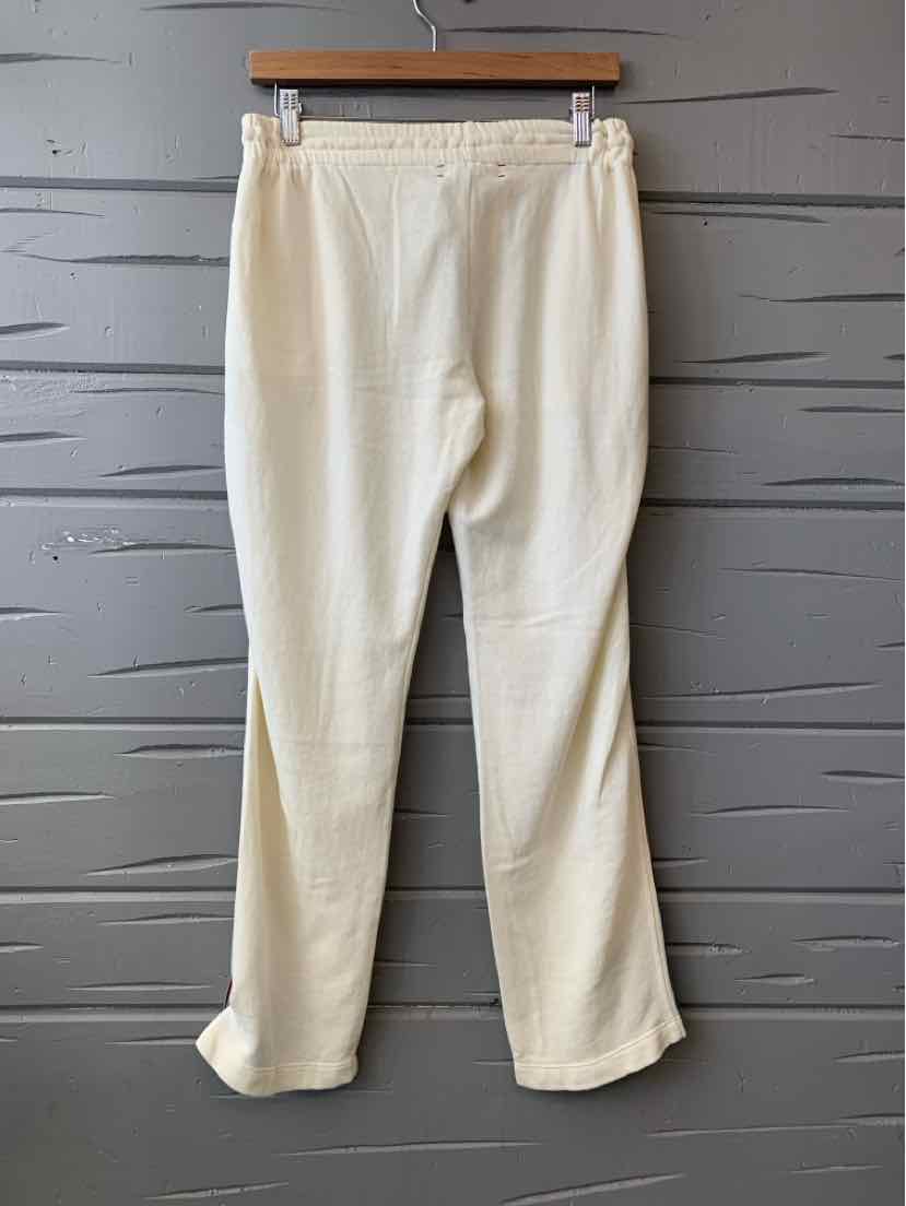 Cream-colored relaxed fit pants with elastic waistband and straight legs hanging on wooden hanger against gray slatted wall.