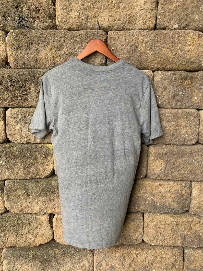 Light gray short-sleeve t-shirt in small size, soft fabric with crew neckline, hanging on wooden hanger against stone wall.