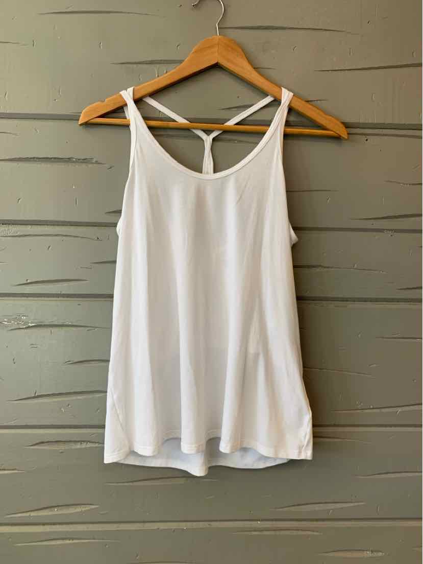 White sleeveless tank top with thin straps and racerback detail hanging on wooden hanger against gray wooden wall.