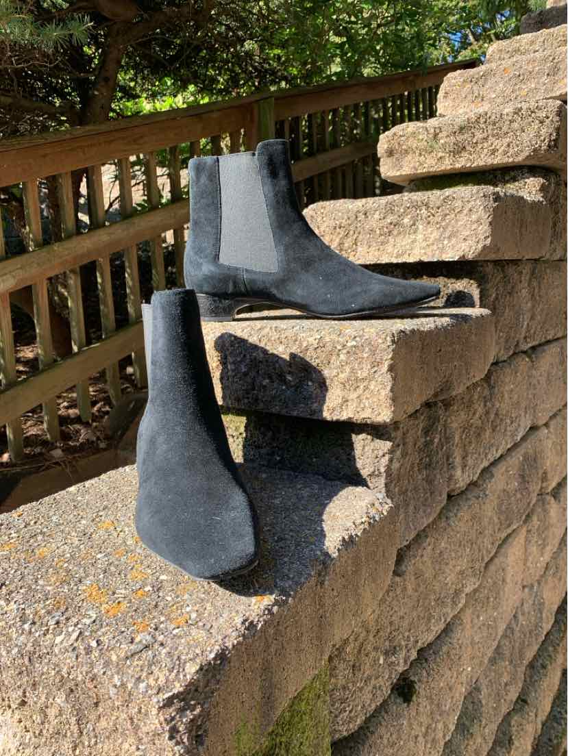 Black suede ankle boots with pointed toes, low heels, and sleek pull-on shafts displayed on a stone wall.