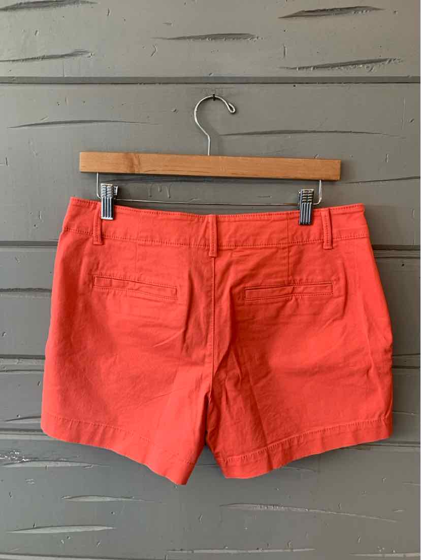 Coral-colored cotton shorts with front pockets and belt loops hanging on wooden hanger against gray wood panel background.