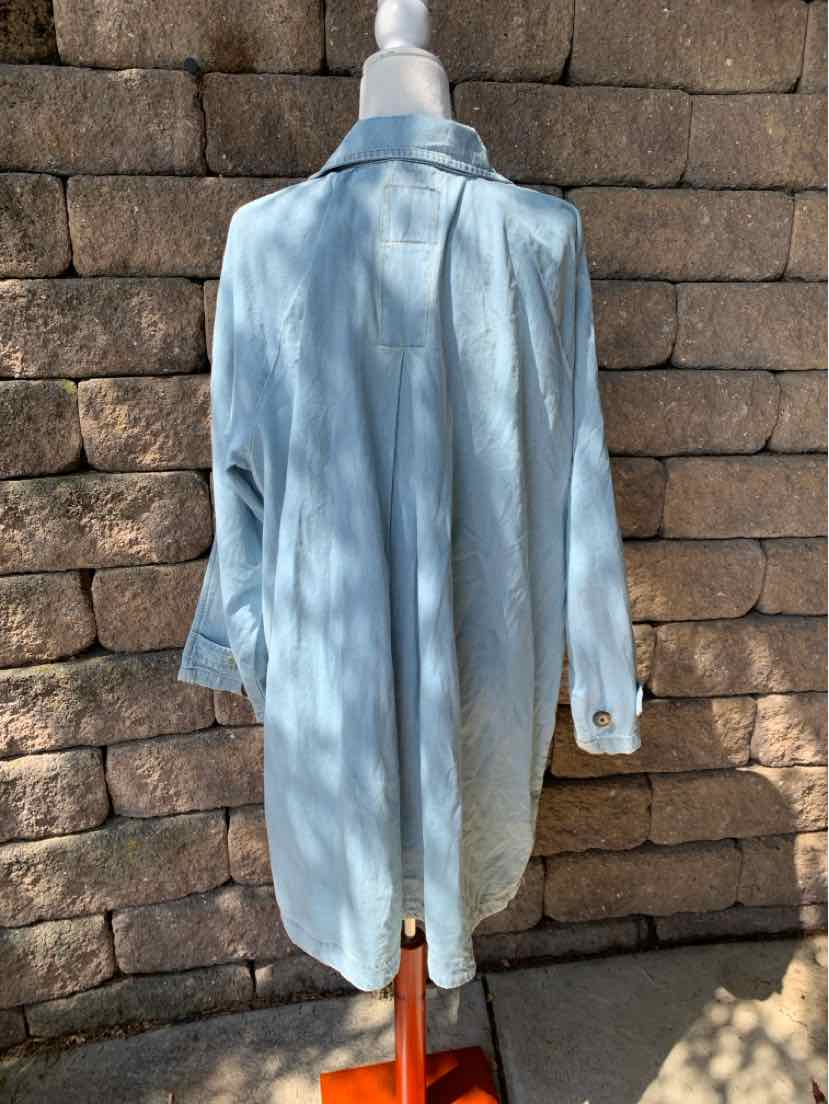 Light blue long-sleeve shirt with buttoned cuffs and a longer back hem on a mannequin against a brick wall.