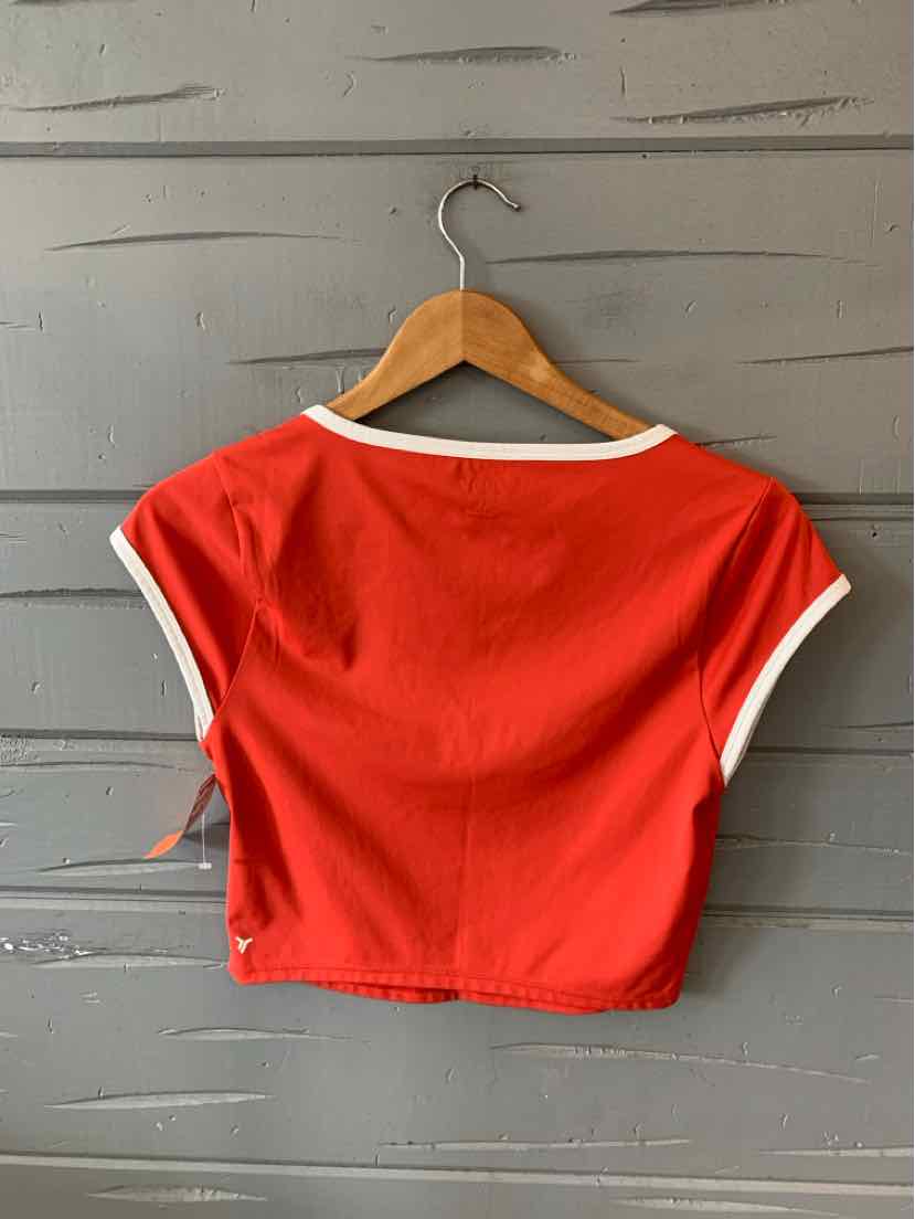 Red cropped t-shirt with white trim on sleeves and neckline hanging on wooden hanger against gray wall.