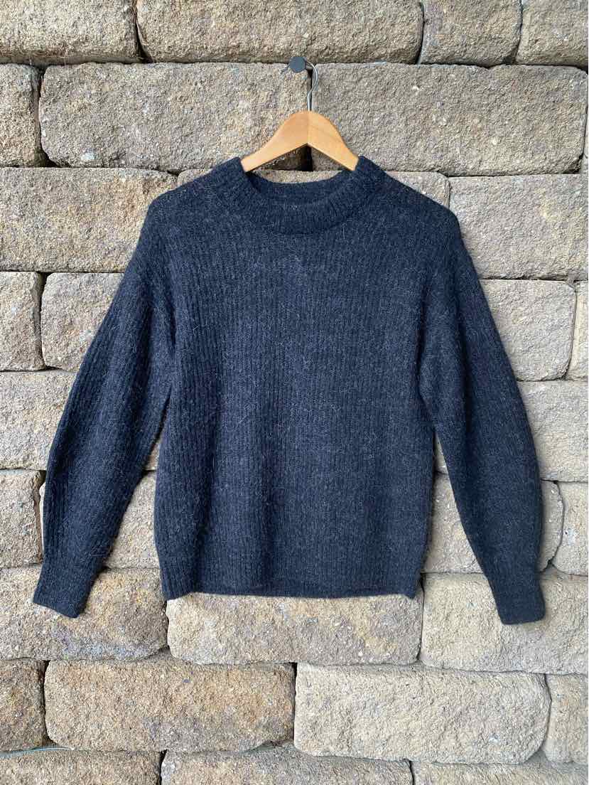 Dark gray knit sweater with round neckline and long sleeves hanging on wooden hanger against stone wall background.