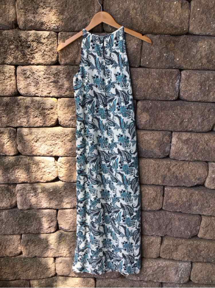 Sleeveless blue paisley maxi dress with gathered neckline hanging on wooden hanger against stone wall.